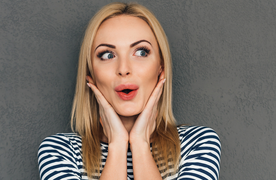 Woman with smooth skin and full, red lips looks surprised with hands on either side of face